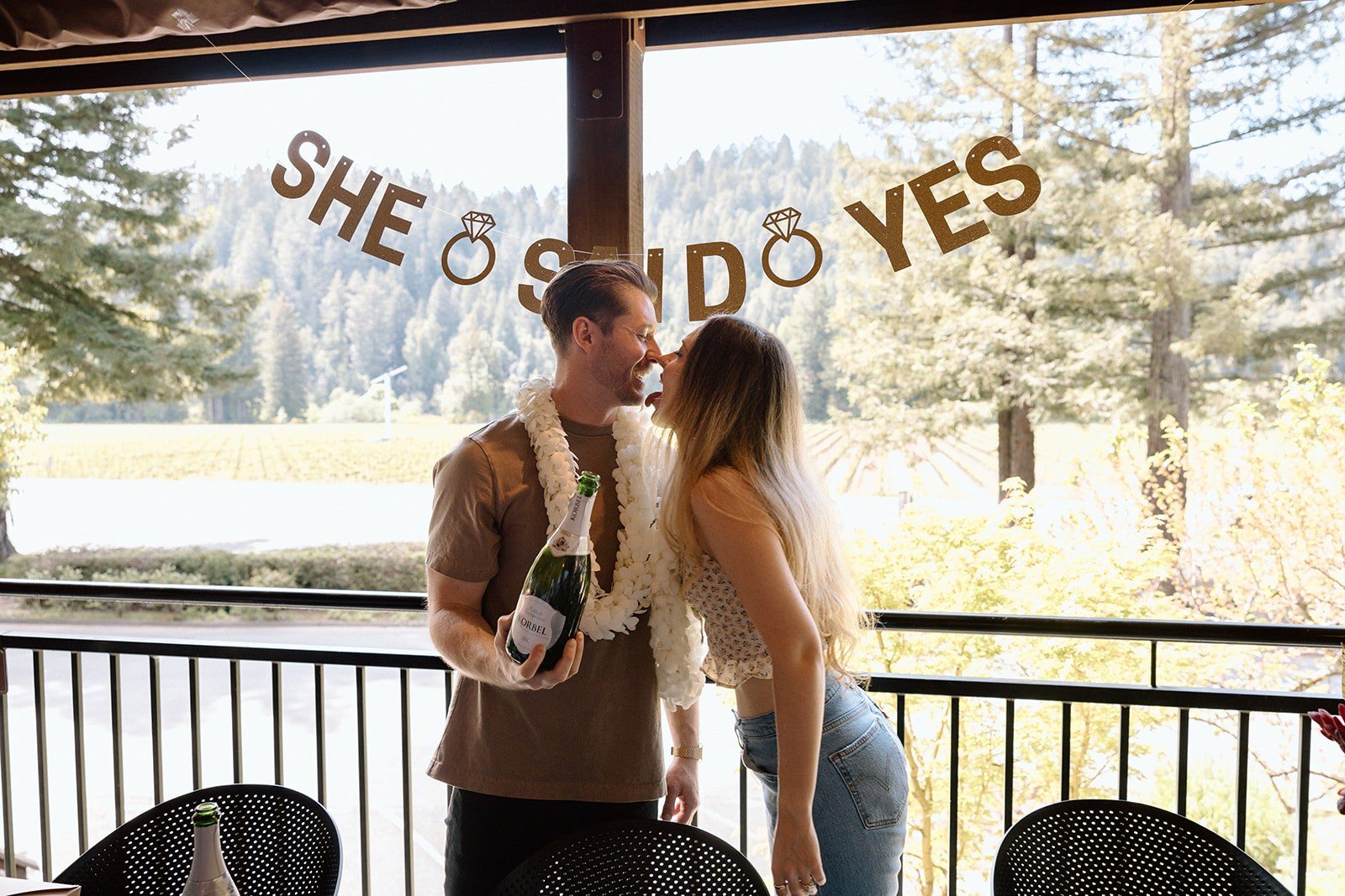 Abel and Aidan kissing under a She Said Yes banner at Korbel, bottle of Korbel in hand