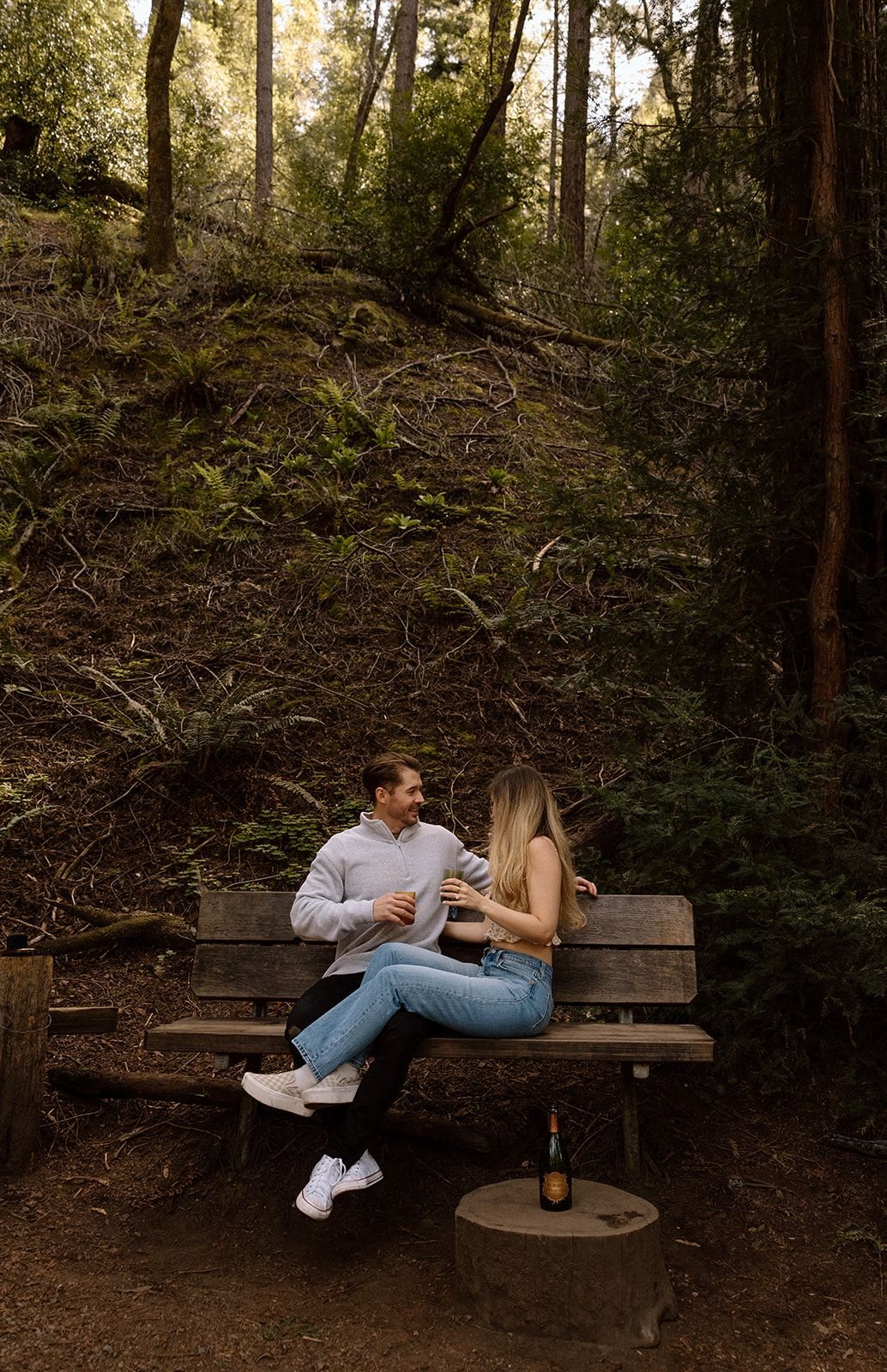 Abel and Aidan sharing a drink on the bench in the redwoods, a bottle of Korbel on the stump beside them